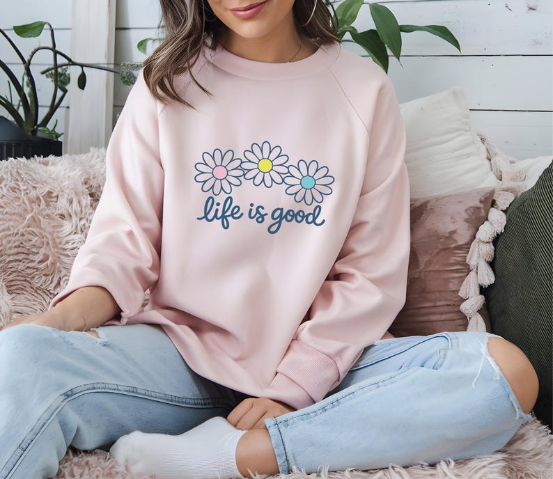 May include: A pale pink sweatshirt featuring a daisy design and the words "life is good". The daisy graphic has pink, yellow, and blue centres. The sweatshirt is a casual, long-sleeved top, suitable for everyday wear.