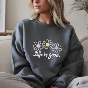 May include: A dark grey sweatshirt featuring a daisy graphic and the words "life is good" in white script. The daisy design has pink, yellow, and blue centres. The sweatshirt is a casual, comfortable style.