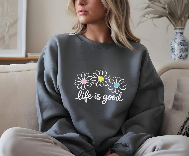 May include: A dark grey sweatshirt featuring a daisy graphic and the words "life is good" in white script. The daisy design has pink, yellow, and blue centres. The sweatshirt is a casual, comfortable style.