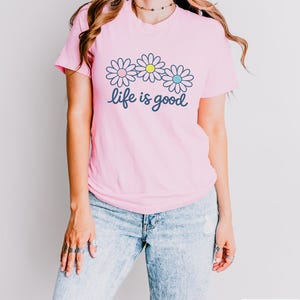 May include: A pink t-shirt featuring a daisy design and the words "life is good". The daisies have white petals with blue, yellow, and blue centres. The model is wearing light blue jeans.
