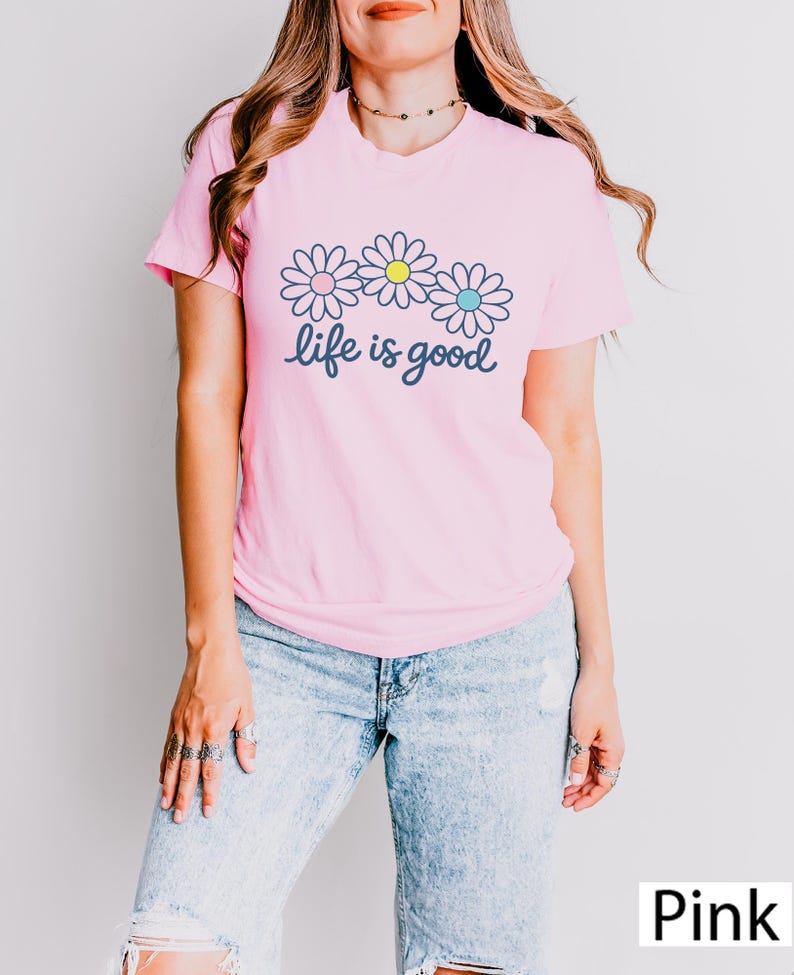 May include: A pink t-shirt featuring a daisy design and the words "life is good". The daisies have white petals with blue, yellow, and blue centres. The model is wearing light blue jeans.