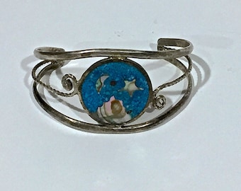 May include: A silver bracelet with a blue and white circular inlay. The inlay features a floral design with a turquoise background and white accents.