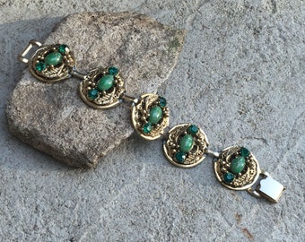May include: A gold tone bracelet with five green oval stones surrounded by smaller green rhinestones. The bracelet has a clasp closure.