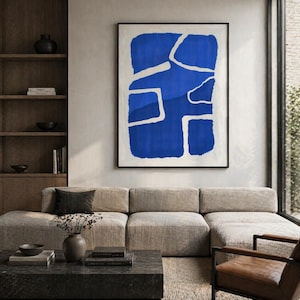 Puede incluir: Gran obra de arte abstracto con un diseño azul audaz sobre fondo blanco, enmarcada en negro. La obra cuelga sobre un sofá seccional beige en un salón moderno. Una mesa de centro gris oscuro y una silla de cuero marrón completan la escena.
