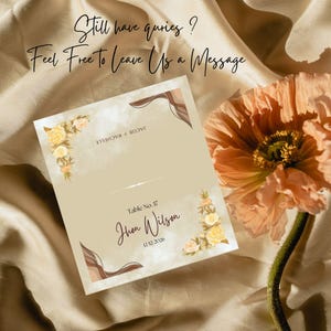 May include: A beige wedding table card with floral accents and the text "Jhon Wilson" and "12/12/2026". The card also includes the text "Still have queries? Feel Free To Leave Us a Message". A peach flower is in the background.