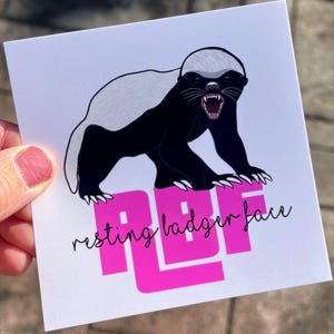 May include: A white card with a black and white illustration of a honey badger with its mouth open. The text "resting badger face" is written in black below the badger. The text "RBF" is written in pink in large letters.