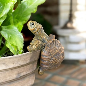 May include: A detailed, bronze-coloured turtle figurine, perched on the edge of a light-coloured planter. The turtle's head and shell are intricately textured, with a detailed eye. The figurine adds a touch of nature-inspired decor to any garden or patio.