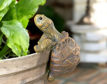 Puede incluir: Una detallada figura de tortuga de color bronce, posada en el borde de una maceta de color claro. La cabeza y el caparazón de la tortuga están intrincadamente texturizados, con un ojo detallado. La figura añade un toque de decoración inspirada en la naturaleza.