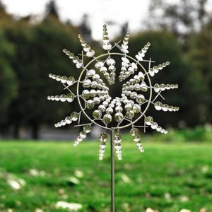 May include: A silver metal wind spinner with a circular design and multiple curved blades. The blades are adorned with small, reflective spheres. The spinner is mounted on a slender metal pole, set against a backdrop of green grass and trees.