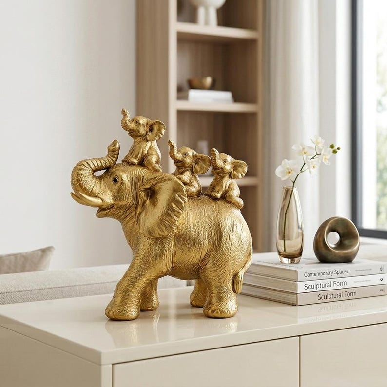 May include: A gold-colored decorative figurine featuring a large elephant with its trunk raised, carrying three smaller elephant calves on its back. The sculpture has a textured surface and is crafted from a metallic material with a polished finish.