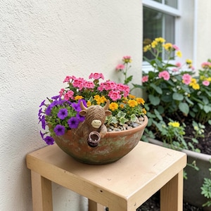 Puede incluir: Una maceta de terracota llena de vibrantes flores rosas, amarillas y moradas. Una figura decorativa de vaca de las Tierras Altas está colocada entre las flores, sosteniendo un pequeño vaso. La maceta está sobre una mesa de madera clara.