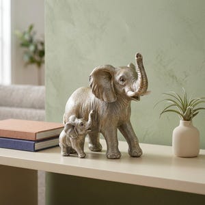 May include: A metallic silver-toned sculpture depicting a large adult elephant standing beside a smaller calf. The adult elephant has its trunk raised upward, and both figures feature textured skin details and smooth, light-colored tusks.