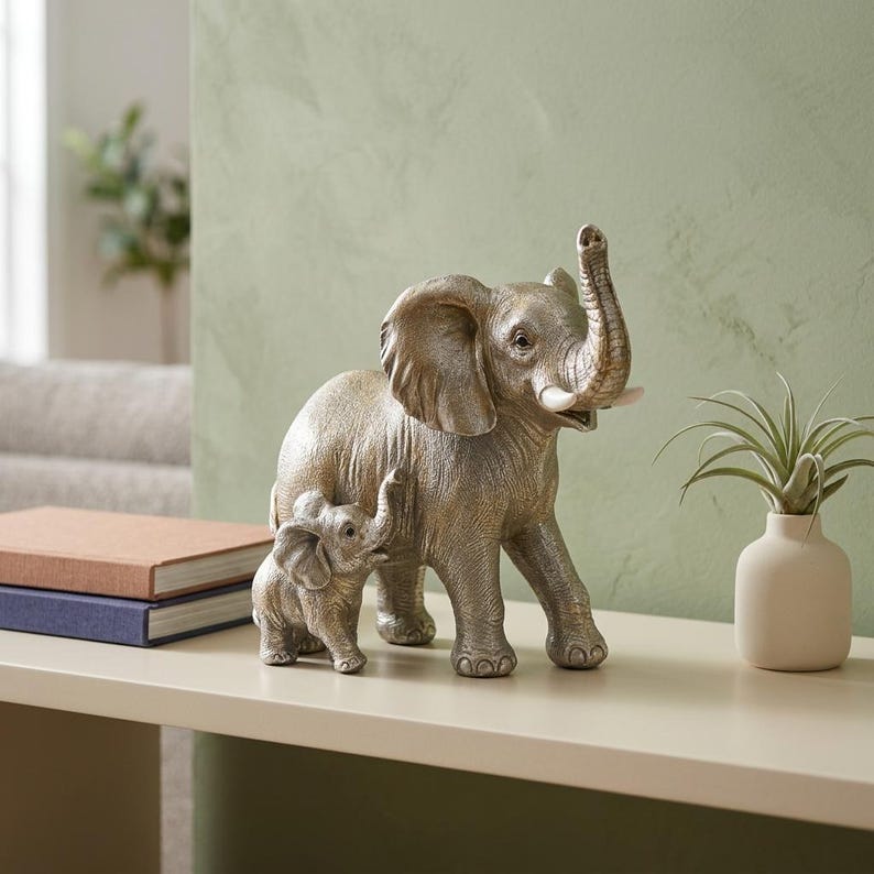 May include: A metallic silver-toned sculpture depicting a large adult elephant standing beside a smaller calf. The adult elephant has its trunk raised upward, and both figures feature textured skin details and smooth, light-colored tusks.