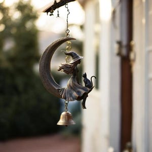 May include: A bronze-toned wind chime featuring a witch silhouette seated on a crescent moon with a black cat. The witch wears a pointed hat and flowing dress. A small bell hangs below the moon. The background is blurred, suggesting an outdoor setting.