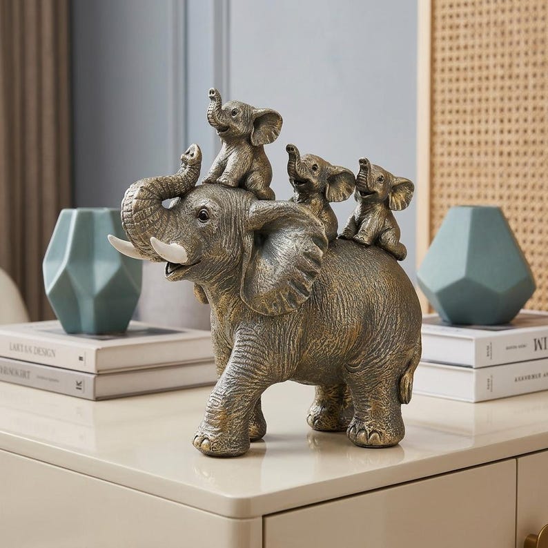 May include: A bronze-toned decorative sculpture featuring a large adult elephant with its trunk raised, carrying three smaller baby elephants on its back. The adult elephant has prominent white tusks and a textured, weathered finish throughout.
