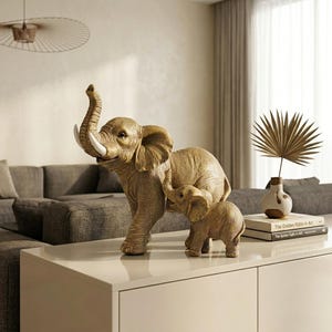 May include: A gold-toned sculpture of an adult elephant with its trunk raised and a smaller calf standing beside it. The figures feature textured skin and white tusks, capturing a realistic depiction of the animals in a standing position.
