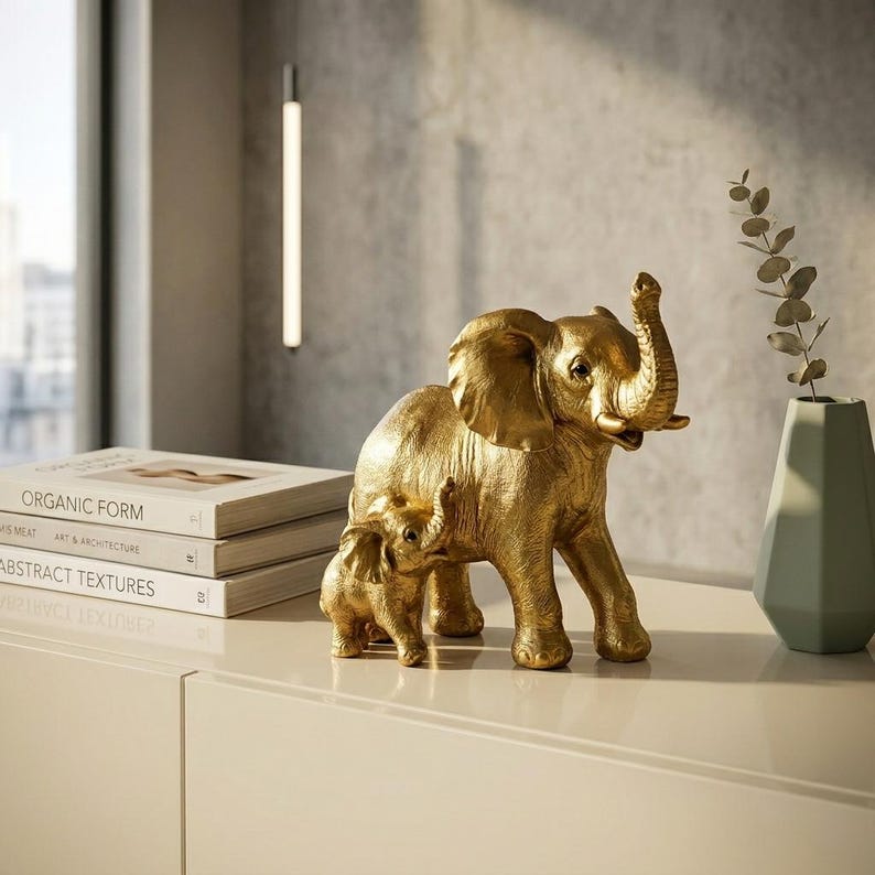 May include: A gold-colored sculpture of a large elephant standing beside a smaller elephant. Both figures feature textured skin and are depicted with their trunks raised. The sculpture is positioned on a smooth surface next to a stack of books.