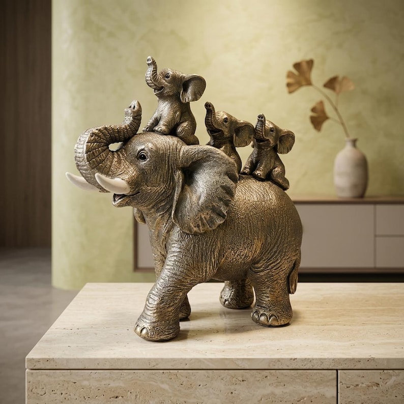 May include: A bronze-toned sculpture of a large elephant with its trunk raised, carrying three smaller elephant figurines on its back. The large elephant features textured skin and smooth, off-white tusks, while the smaller elephants are positioned in a row along its spine.