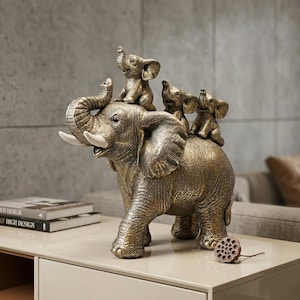 May include: A bronze-colored decorative figurine featuring a large adult elephant with its trunk raised, carrying three smaller baby elephants on its back. The adult elephant has prominent white tusks and textured skin, while the smaller elephants are depicted in seated positions.