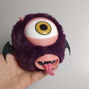 May include: A plush, purple monster toy with a large, white and yellow eye, pink eyelids, and a pink tongue. The toy has small horns, black wings, and a wide open mouth. The toy is held in a hand.
