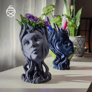 Könnte beinhalten: Zwei 3D-gedruckte, kopfförmige Pflanzgefäße mit fließenden Haarstrukturen. Das vordere Gefäß ist mattgrau und enthält violette Kunstblumen. Das hintere Gefäß ist dunkelblau mit glitzerndem Finish und dient als Halter für Stifte.