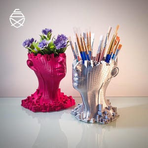 Peut inclure: Deux récipients en forme de tête avec une texture pixélisée et cubique. Le récipient de gauche est magenta et contient des fleurs artificielles violettes. Celui de droite est argenté métallique et contient divers pinceaux à manche en bois.