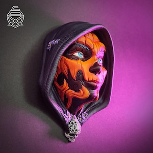 May include: A wall-mounted sculpture featuring a face with orange pumpkin-like skin and bright blue eyes, framed by a dark grey hooded cloak. The hood is decorated with small purple flowers and a detailed white skull charm at the bottom center.