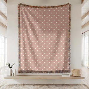 May include: A woven throw blanket in a dusty rose pink color featuring a repeating pattern of small white polka dots. The blanket has a textured appearance with decorative fringe along the top and bottom edges, showcasing multicolored threads.