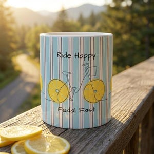 May include: A ceramic mug featuring a light blue and white vertical striped pattern. The design includes a black line drawing of a bicycle with yellow lemon slices as wheels. The text "Ride Happy" and "Pedal Fast" is printed in black lettering.