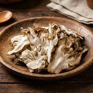 Può includere: Una ciotola di legno piena di funghi secchi di colore chiaro. I funghi hanno un aspetto strutturato e stratificato e sono disposti a cerchio. La ciotola è appoggiata su una superficie di legno rustica, con un cucchiaio di legno e un panno di lino sullo sfondo.