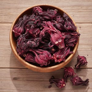 Pode incluir: Uma tigela de madeira cheia de flores de hibisco secas. As flores são de uma cor bordô profunda com uma aparência seca e texturizada. Algumas flores estão espalhadas fora da tigela sobre uma superfície de madeira marrom clara.