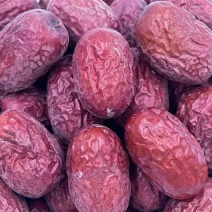 Puede incluir: Primer plano de un montón de dátiles rojos frescos. Los dátiles tienen una textura arrugada y forma ovalada. El color varía de rojo intenso a un tono más claro, lo que sugiere diferentes grados de madurez. La imagen está bien iluminada, destacando los detalles.