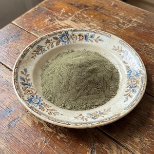 以下が含まれることがあります： 青と茶色の花柄模様の入った、オフホワイトのヴィンテージセラミックプレートに、細かい緑色の粉末が盛られています。 プレートは、古びた木製の表面に置かれ、素朴な雰囲気を醸し出しています。