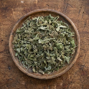 Puede incluir: Un cuenco de madera lleno de hierbas verdes secas. Las hierbas tienen un aspecto texturizado y son una mezcla de verde claro y oscuro. El cuenco está sobre una superficie de madera rústica.