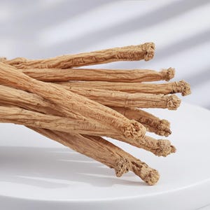 May include: A close-up of several dried ginseng roots. The roots are a light brown color with a textured, wrinkled surface. The ends of the roots have a knobby appearance. The roots are arranged on a white surface.