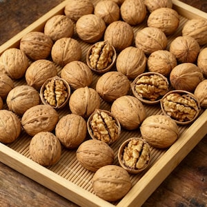 May include: A wooden tray filled with whole walnuts and a few opened to reveal the nutmeat. The walnuts are brown and have a textured shell. The tray sits on a wooden surface.