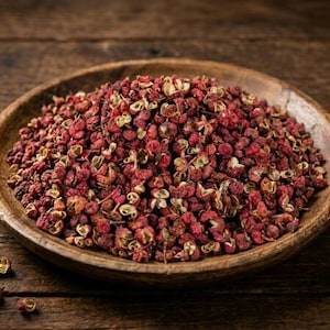 Pode incluir: Um monte de grãos de pimenta de Sichuan vermelhos secos empilhados numa tigela de madeira circular e rasa. Os grãos de pimenta têm uma aparência texturizada e descascada com tons de vermelho escuro e castanho, com alguns grãos espalhados.