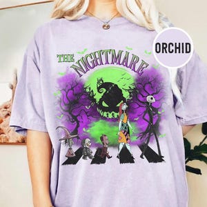 May include: A light purple t-shirt featuring a graphic from "The Nightmare Before Christmas." The design includes Jack Skellington, Sally, and other characters crossing a street, with a green and purple background. The text "THE NIGHTMARE" is arched above the scene.