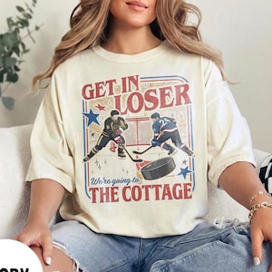May include: Ivory-colored t-shirt with a hockey-themed graphic. The design features two hockey players, a puck, and the text "Get In Loser, We're going to The Cottage" in red and blue. The shirt is a relaxed fit.
