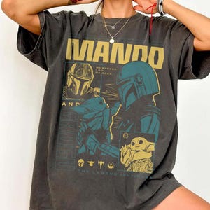May include: Dark grey t-shirt with a gold and teal graphic design. The design features the word "MANDO", images of a Mandalorian helmet and Grogu, and the phrases "WHEREVER I GO HE GOES" and "THE LEGEND CONTINUES".