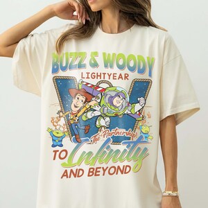 May include: Cream-colored t-shirt featuring a graphic of Buzz Lightyear and Woody from Toy Story. The design includes the text "Buzz & Woody Lightyear" and "To Infinity and Beyond" in green and blue. The shirt has short sleeves and a relaxed fit.