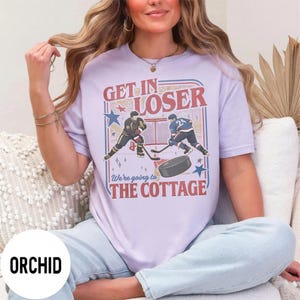 May include: A light purple t-shirt with a hockey-themed graphic. The graphic features two hockey players, a net, a puck, and the text "GET IN LOSER" and "We're going to THE COTTAGE" in red and blue. The shirt is a casual wear item.