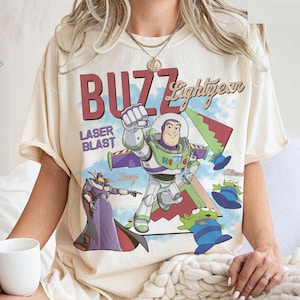 May include: A cream-colored short-sleeve t-shirt featuring a graphic print of Buzz Lightyear, Zurg, and aliens. The text reads BUZZ Lightyear and LASER BLAST. The design includes a blue sky background with geometric shapes in red and green.
