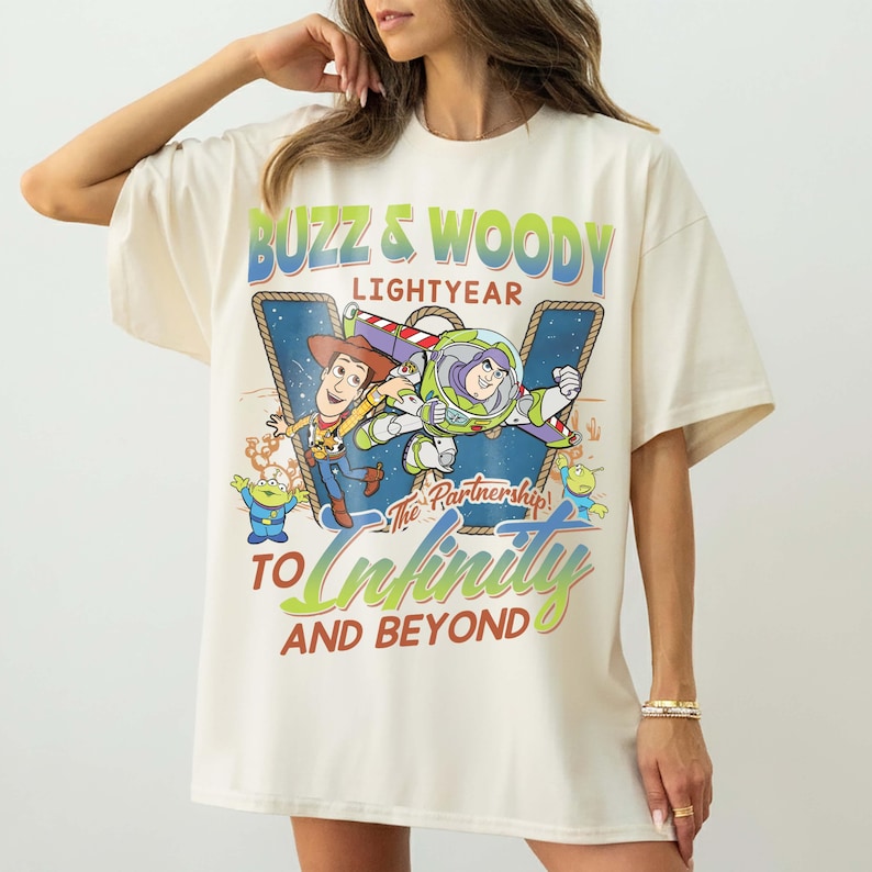 May include: Cream-colored t-shirt featuring a graphic of Buzz Lightyear and Woody from Toy Story. The design includes the text "Buzz & Woody Lightyear" and "To Infinity and Beyond" in green and blue. The shirt has short sleeves and a relaxed fit.