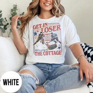 May include: White t-shirt with a hockey-themed graphic. The design features two hockey players, a puck, and the text "GET IN LOSER" and "We're going to THE COTTAGE" in red and blue.