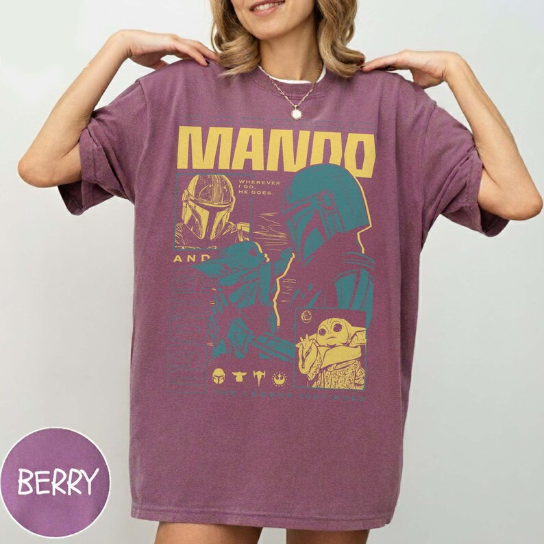 May include: Berry-coloured t-shirt with a graphic design featuring the word "MANDO" in yellow block letters. The design includes illustrations of characters from a popular science fiction series. The shirt has short sleeves and a relaxed fit.