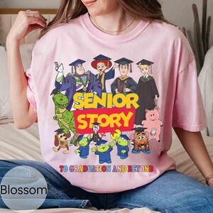 K&ouml;nnte beinhalten: Hellrosa T-Shirt mit einer Grafik von Zeichentrickfiguren in Abschlusskleidung. Der Text "SENIOR STORY" ist in Gelb und Rot gehalten, darunter der Schriftzug "TO GRADUATION AND BEYOND".