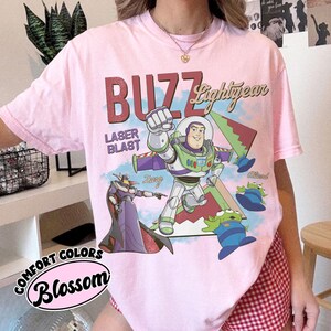 May include: A light pink t-shirt featuring a graphic print of Buzz Lightyear, Zurg, and three green aliens. The text reads BUZZ Lightyear, LASER BLAST, Zurg, and Alien. The shirt has a crew neckline and short sleeves, with a Comfort Colors Blossom label.
