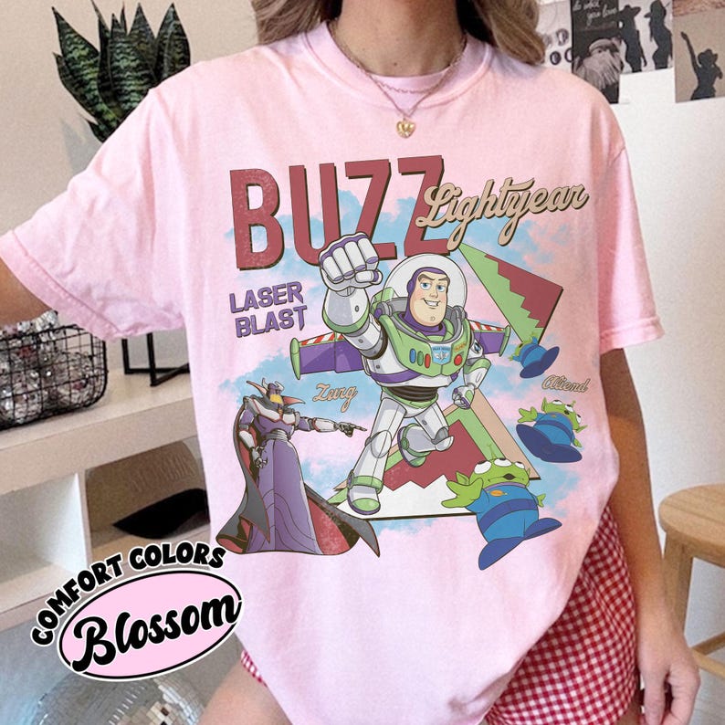 May include: A light pink t-shirt featuring a graphic print of Buzz Lightyear, Zurg, and three green aliens. The text reads BUZZ Lightyear, LASER BLAST, Zurg, and Alien. The shirt has a crew neckline and short sleeves, with a Comfort Colors Blossom label.