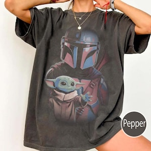 May include: A charcoal gray short-sleeve t-shirt featuring a graphic print of a helmeted warrior in red and blue armor standing behind a small green creature with large ears. The word Pepper is printed in white on a small circular badge.